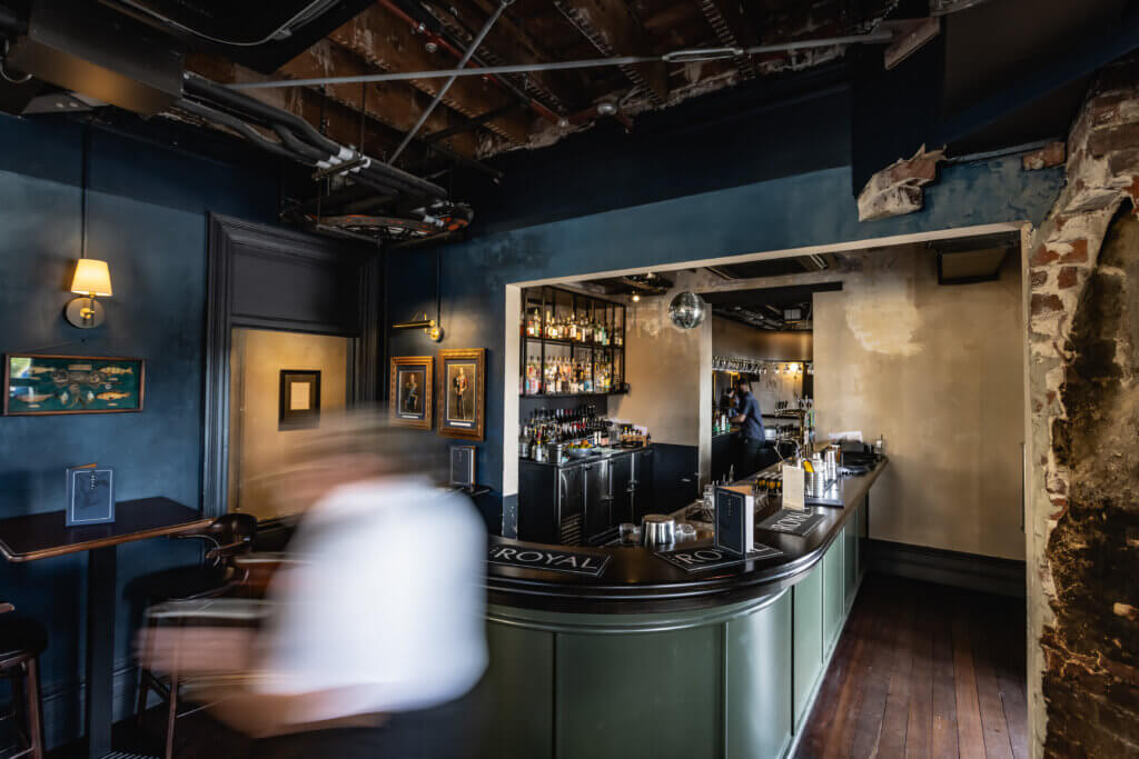 Busy boutique bar with curved counter, staff serving drinks and exposed ceiling at The Royal.
