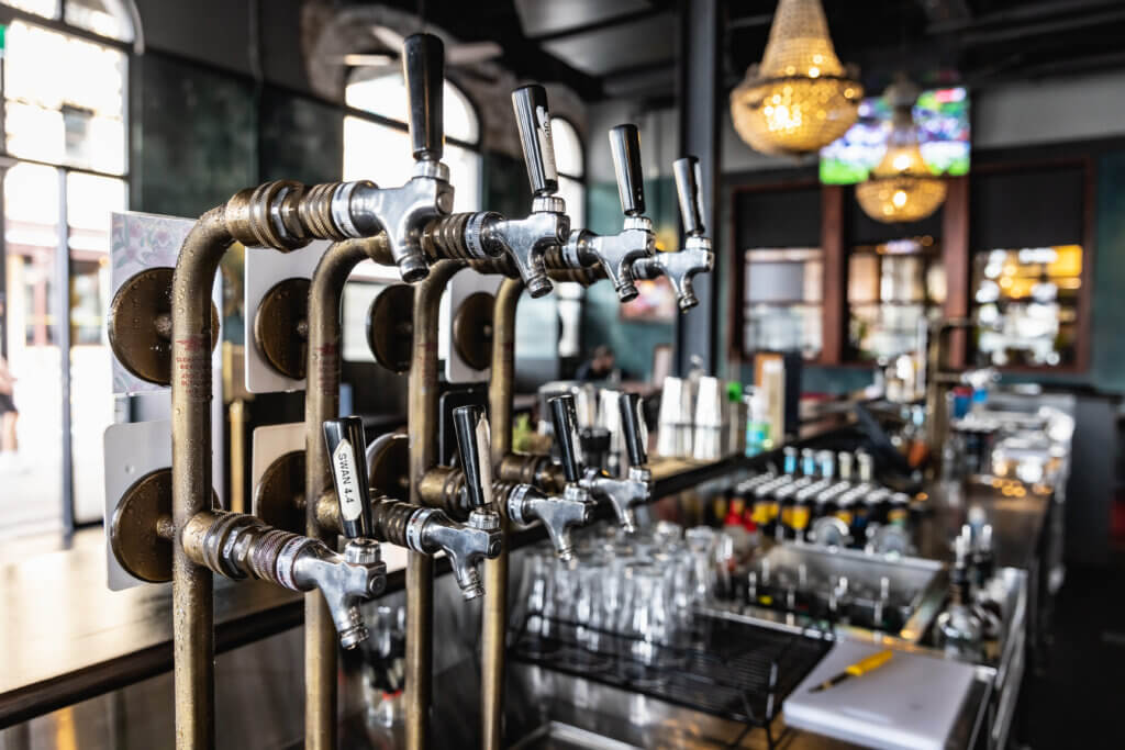 Multiple stainless steel beer taps on a vintage-style bar system with glassware, spirits and chandeliers in background.