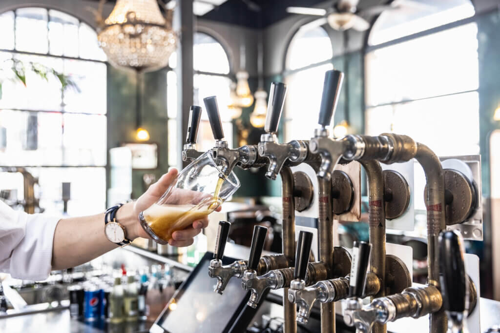 Bartender pouring a foamy beer from a multi-tap system in a bright bar with chandeliers and arched windows.