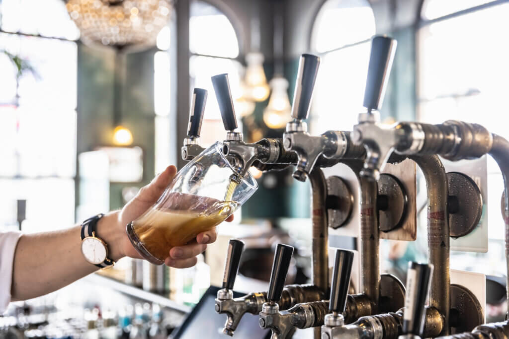 Bartender tilting a glass while pouring foamy beer from a tap system in a bright, elegant bar setting.