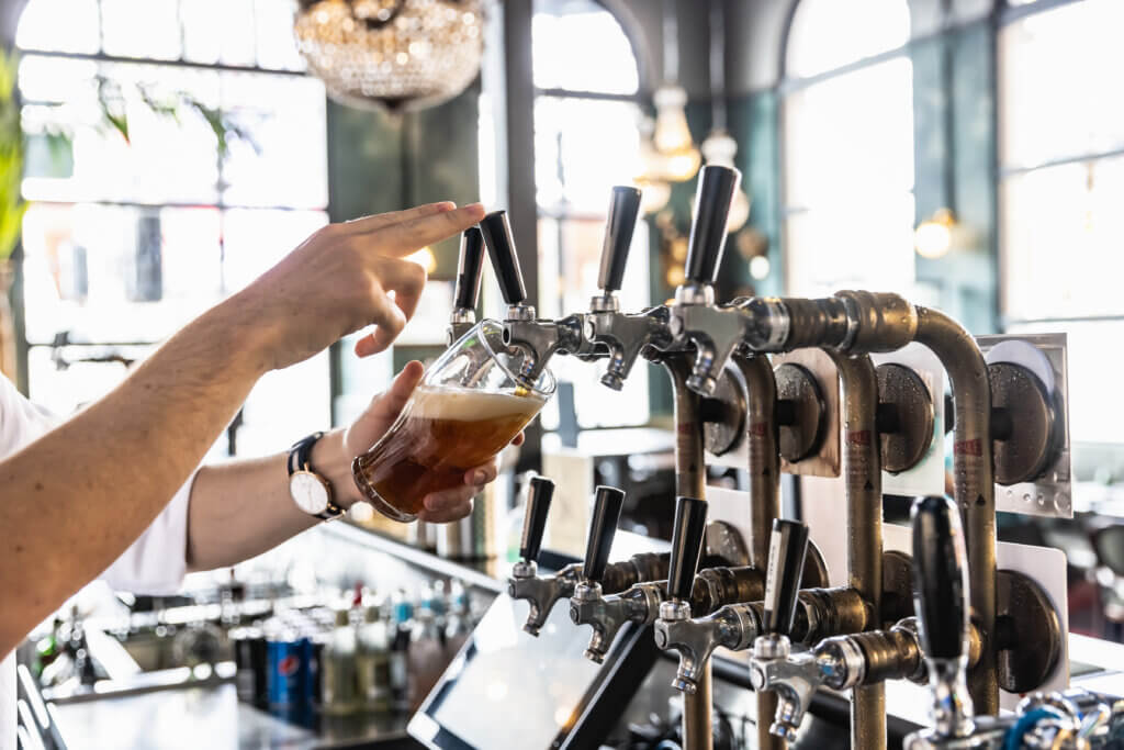Bartender pouring amber beer from a vintage tap system at a well-lit bar with chandeliers and tall windows.