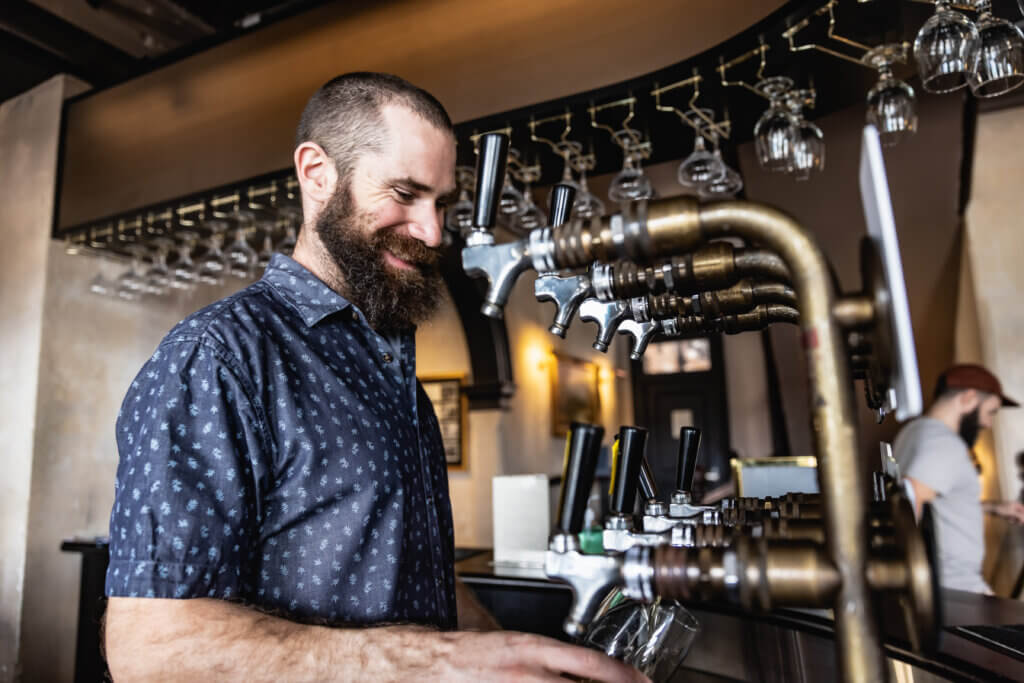 Smiling bartender using a vintage-style beer tap system with hanging glassware overhead.