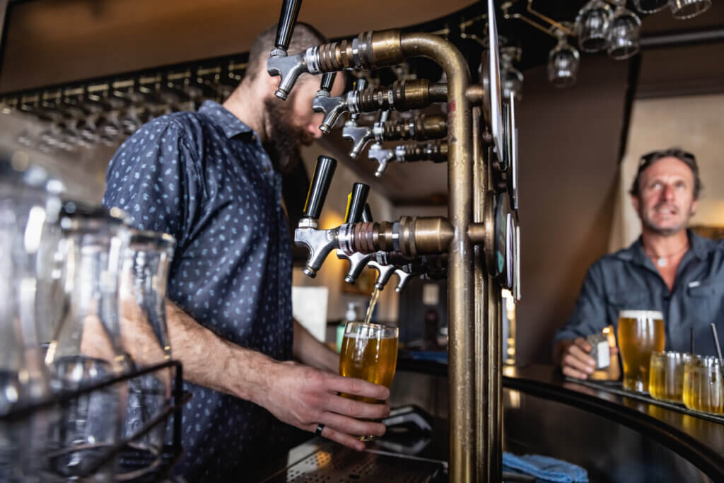 Bartender pouring a beer from a vintage-style tap system while a patron waits at the bar with a drink.