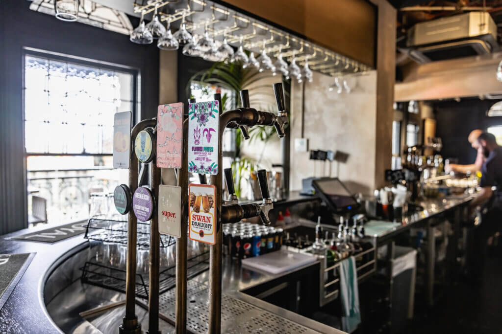 Close-up of beer taps with colourful labels at a modern bar, with glassware and staff in the background.