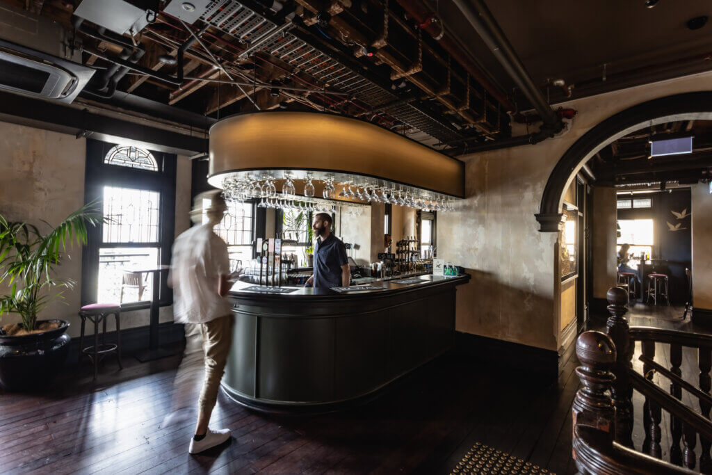Stylish heritage bar with curved counter, overhead glass rack and tap beer system, with patrons and staff in view.