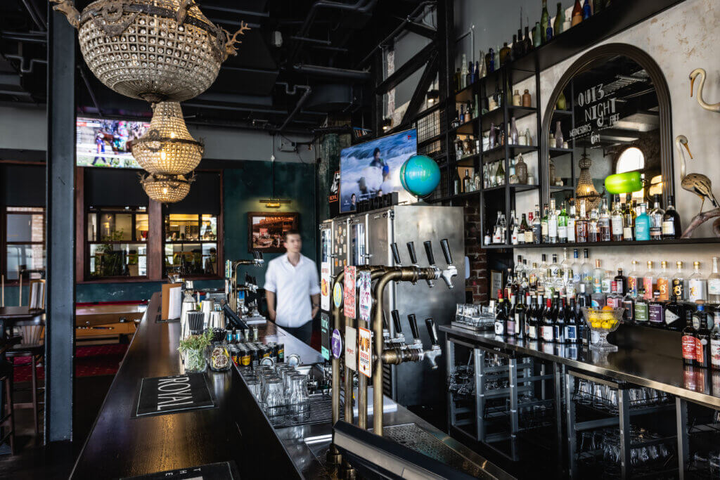 Beer taps with branded handles behind modern bar counter, with spirits display and chandeliers overhead.