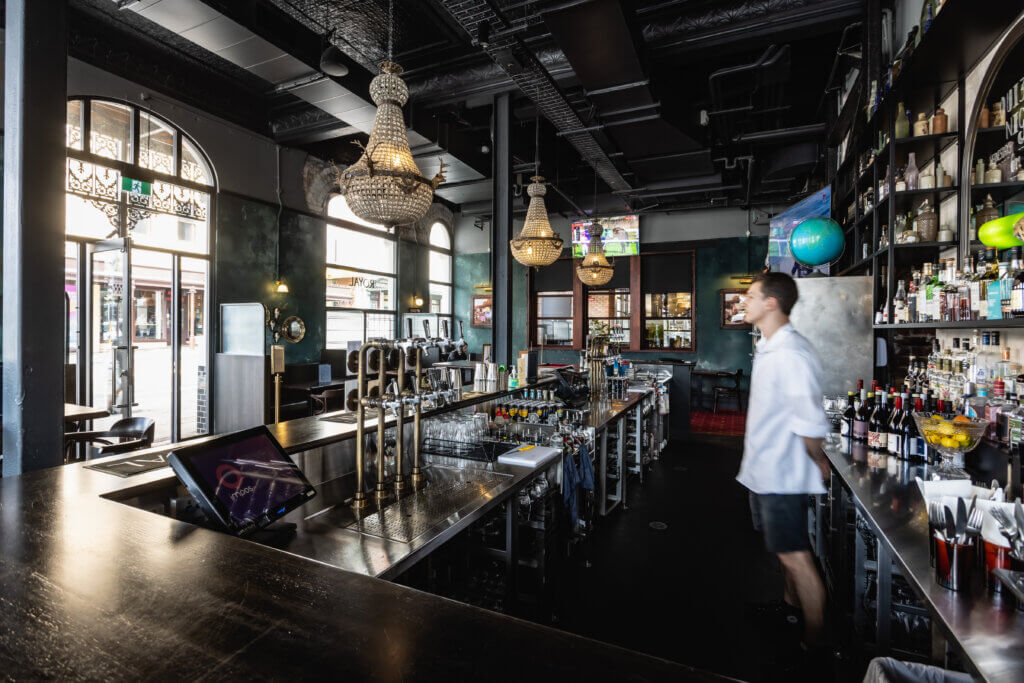 Bartender behind modern stainless steel bar with beer taps, beverage displays and chandeliers in stylish venue.
