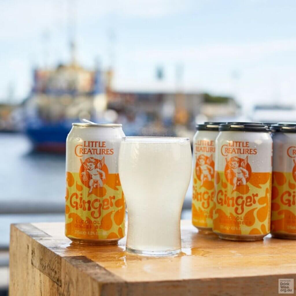 Little Creatures Ginger Beer cans and glass on wooden table with waterfront backdrop.