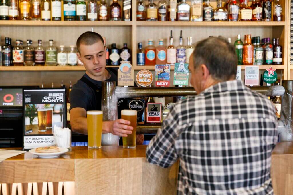 Bartender serving pints of beer to customer at The Elford bar, with spirits displayed on shelves behind.