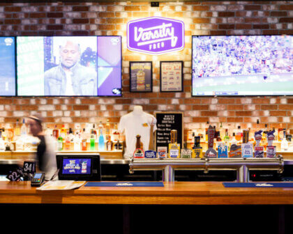 Varsity bar interior with branded beer taps, TVs playing sport, and exposed brick wall backdrop.