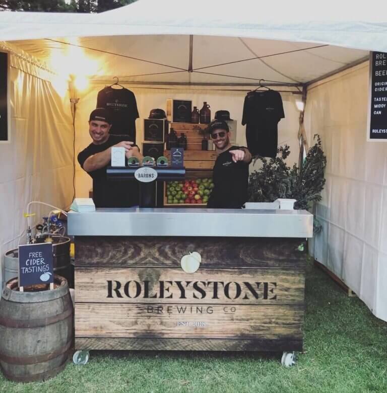 Two staff serving cider at a Roleystone Brewing Co. market stall with Barons tap system and free tastings sign.