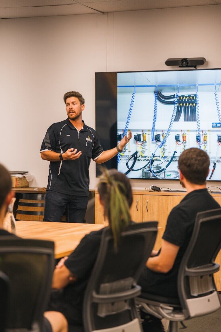 Man in Barons uniform giving technical training to staff, with beverage system setup on screen.